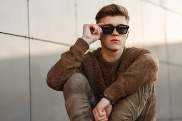 Wall Mural - handsome fashionable young man in sunglasses with a knitted sweater and fashion military pants sits on the street