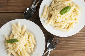 Cooked penne pasta with butter and cheese, isolated in white round plates, over wooden table, decorated with basil leaves. Top view.