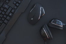 Black Mouse, The Keyboard, The Headphones Are Isolated On A Dark Background, The Top View. Flat Lay Gamer Background. Workplace With A Keyboard, Mouse And Headphones On A Black Background. Copyspace