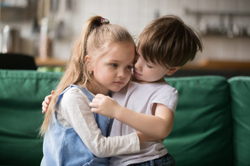 little boy hugging consoling upset girl sitting on sofa, kid brother embracing sad sister apologizin
