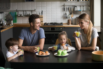 Happy family and kids having breakfast sitting at kitchen table, smiling mom and dad eating together with children talking enjoying tasty food fresh juice and cakes at home, morning lifestyle concept