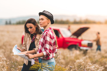 Wall Mural - serious young girlfriends with map standing in field and trying to navigate