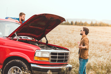 Wall Mural - young men standing near car with broken engine in field