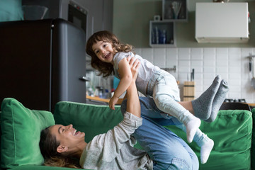 Young mother playing with cute baby girl on sofa at home, happy single mom or sister laughing lifting little kid daughter lying on sofa on weekend, family mum and child having fun together concept