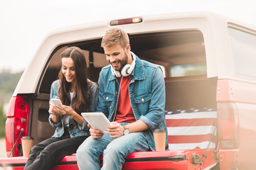 Wall Mural - happy young couple using gadgets while sitting in car trunk during trip