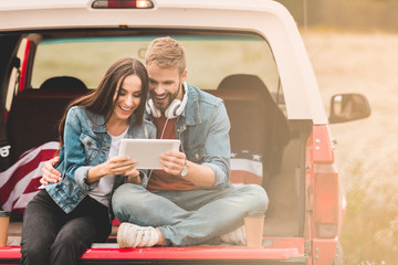 Wall Mural - beautiful young couple using tablet while sitting in car trunk during trip