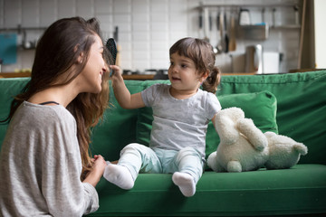 Wall Mural - Cute kid girl brushing mothers hair having fun together on sofa, little daughter holding brush helping young happy single mom with hairstyle playing at home, child and mommy funny activity concept