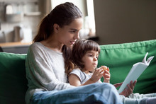 Young Mother Teaching Daughter Holding Book Sitting On Sofa, Mom Or Babysitter Embracing Little Girl Reading Fairy Tale To Kid, Nanny With Child Having Fun Together At Home, Family Leisure Activities