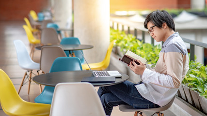 Wall Mural - Young Asian student man with eyeglasses dressed in casual style reading book and using laptop computer in college building. Education research and self studying in university life concepts