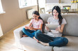 © estradaanton - Woman and her daughter are sitting on sofa with their legs crossed and reading. Woman holds tablet while girl has phone. She smiles.
