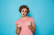 Portrait of stylish handsome man 20s with brown curly hair wearing modern glasses pointing fingers at camera, isolated over blue background