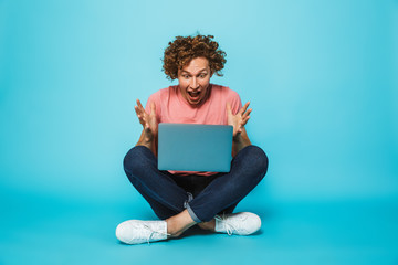 Poster - Photo of surprised or shocked guy 20s with brown curly hair looking at silver laptop, while sitting on floor with legs crossed, isolated over blue background