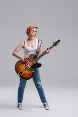 Wall Mural - Young woman musician with an acoustic guitar in hand on a gray background. He laughs and plays rock and roll loudly. Full-length portrait