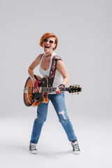 Wall Mural - Young woman musician with an acoustic guitar in hand on a gray background. He laughs and plays rock and roll loudly. Full-length portrait