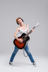 Wall Mural - Young woman musician with an acoustic   guitar in hand and in headphones on a gray background. He laughs and plays rock and roll loudly. Full-length portrait