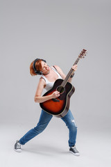 Wall Mural - Young woman musician with an acoustic   guitar in hand and in headphones on a gray background. He laughs and plays rock and roll loudly. Full-length portrait