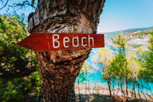 Assos Village In Morning Light, Kefalonia. Greece. Beach Wooden Arrow Sign On A Pine Tree Showing Direction To Small Hidden Beach