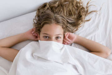 portrait of a young beautiful kid resting on bed, smiling and covering with sheets. white room. Family lifestyle indoors.