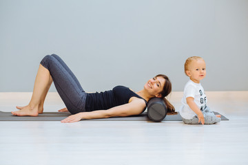 Wall Mural - Young mother relaxing her neck and back with foam roller does physical pilates exercises while her toddler baby son playing near. Fitness, happy maternity sport with children concept.