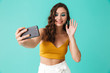 © Drobot Dean - Photo of cheerful charming woman 20s with glamour style smiling and waving hand at camera while taking selfie, isolated over blue background