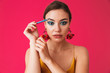 © Drobot Dean - Image of caucasian stylish woman 20s wearing earrings smiling and applying makeup with blue eyeliner, isolated over pink background