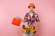© ViDi Studio - Strong young handyman woman in orange helmet, plaid shirt, denim shorts, kit tools belt full of instruments, toolbox isolated on pink background. Female in male work. Renovation and occupation concept