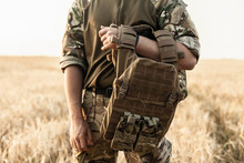Soldier Man Standing Against A Field. Soldier In Military Outfit With Bulletproof Vest. Photo Of A Soldier In Military Outfit Holding A Gun And Bulletproof Vest On Orange Desert Background.