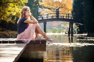 Pretty dreaming young woman sitting on a bridge near lake in autumn park