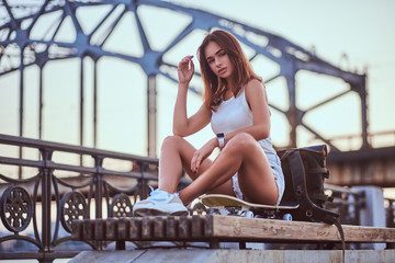 Wall Mural - Young sensual skater girl dressed in shorts and t-shirt sitting on the embankment during beautiful sunset.