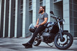 © Fxquadro - Brutal bearded male in a gray t-shirt and black pants holds a helmet sitting on his custom-made retro motorcycle against skyscraper.