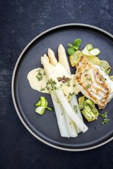 Wall Mural - Modern German fried cod fish filet with white asparagus in hollandaise sauce with roast potatoes and sliced zucchini as top view on a plate with copy space