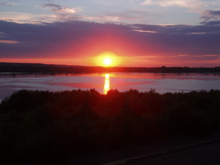 Wall Mural - red-yellow sun setting behind a forest and reflected in a river against a cloudy sky background