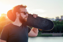Happy Californian Beautiful Bearded Red-haired Sexy Man In Sunflasses Holds A Skateboard (longboard) At Sunset On The Beach. Cool Photosession Photoshoot Of Guy On Summer Park