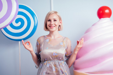 beautiful and young blonde woman sexy model in a chic short dress ,perfect makeup meditating yoga huge lollipop fake sweets сake, candy background in studio. concept of diet and diabetes