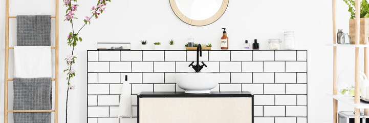 Real photo of white bathroom interior with wooden ladder with towels, tiles on wall shelf with plants and cosmetics and white sink on cabinet