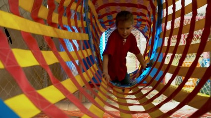 Wall Mural - Front view of playful little girl of elementary school age climbing horizontally through colorful net tunnel in playroom