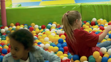 Wall Mural - Follow shot of two little multi ethnic girls having fun in playroom together and sliding down into ball pit