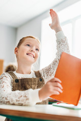 Wall Mural - smiling little schoolgirl with book raising hand during lesson