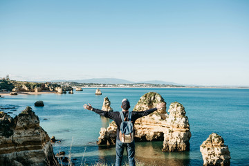 Wall Mural - A tourist or traveler with a backpack admires the beautiful view of the Atlantic Ocean and the coast near the city called Lagos in Portugal and raises his hands up showing how he is free and he likes.