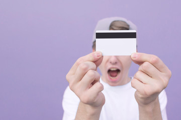 Wall Mural - Emotion young man on a purple background holds a bank card in his hands. Сredit card is in the hands of a young man. Close-up of a bank card