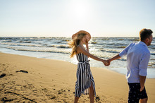 Side View Of Young Smiling Couple In Love Holding Hands On Sandy Beach In Riga, Latvia