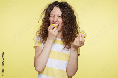 Fotografie, Tablou Portrait of cute Caucasian female with disgusting expression tastes sour lemon, frowns in displeasure, isolated over yellow studio wall background