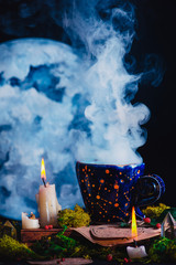 Wall Mural - Blue ceramic cup with constellations and stars in a dark still life. Magician workplace with burning candles, smoke, crystals, moss and potion bottles. Occult concept with copy space