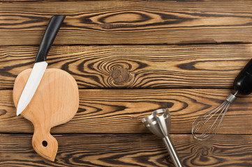 White ceramic knife with black handle and wooden cutting board and whisk and metal blender on old worn brown table. Top view with copy space