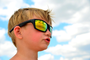 A little boy looks at the cloudy sky with glasses. The clouds are reflected in the glass. Yellow glasses against the blue sky.