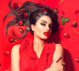 Wall Mural - Beauty sexy model girl lying on red background with rose flowers and strawberries. Beautiful brunette young woman with long hair and perfect make-up, red seductive lips, smoky eyes. Trendy makeup