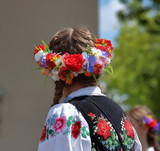 Dziewczyna (górna część ciała) stoi tyłem, włosy uczesane w warkocze, na głowie kolorowy kwietny wianek, ubrana w tradycyjny haftowany strój regionalny