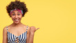 © WHstudio Leushin N - Lovely African American female has gentle pleasant smile, dressed in casual blank and white striped top, has dark skin, points aside with thumb, stands over yellow wall with blank space for you