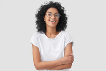 Joyful African American Student Keeps Hands Crossed, Laughs At Good Joke, Wears Casual Clothes And Round Spectacles, Isolated Over White Background. Happy Young Woman With Dark Skin Poses Indoor