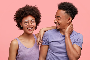 Wall Mural - Photo of joyful dark skinned female and male companions have joy together, dressed casually, smile positively, stand against pink background. Happy African American woman leans at shoulder of man